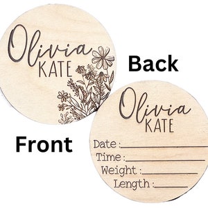 A round wooden disc with a floral design and the name Olivia Kate engraved on the front. The back of the disc has blank lines for recording a baby's date, time, weight, and length. The text on the back reads: Date:, Time:, Weight:, Length:.