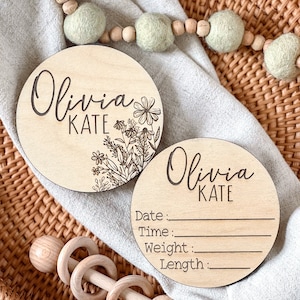 Two round wooden plaques with the name Olivia Kate engraved on them. One plaque has a floral design and the other has blank lines for writing the date, time, weight, and length. The plaques are on a woven wicker surface.