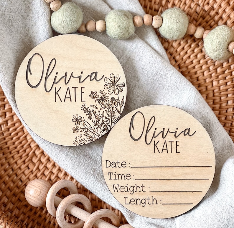 Two round wooden plaques with the name Olivia Kate engraved on them. One plaque has a floral design and the other has blank lines for writing the date, time, weight, and length. The plaques are on a woven wicker surface.