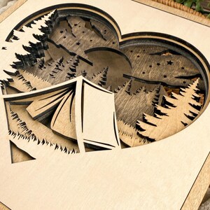 May include: A layered wooden art piece featuring a heart-shaped cutout with a camping scene. The design includes a tent, trees, mountains, a moon, and stars, all in natural wood tones. The artwork is set within a square frame.