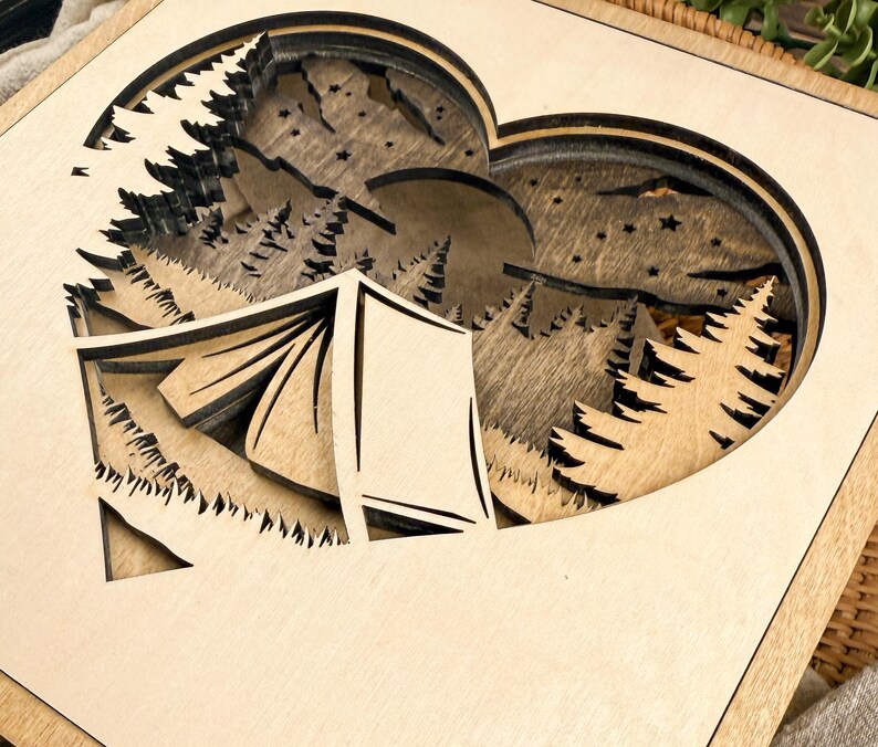 May include: A layered wooden art piece featuring a heart-shaped cutout with a camping scene. The design includes a tent, trees, mountains, a moon, and stars, all in natural wood tones. The artwork is set within a square frame.