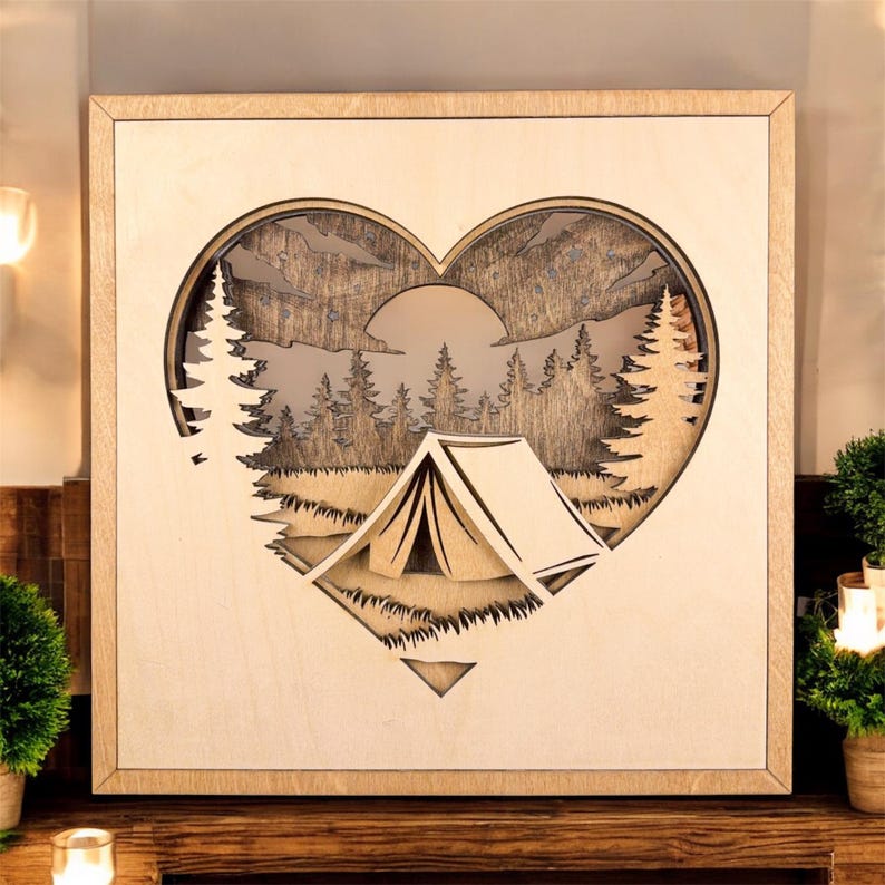 May include: Wooden wall art featuring a heart-shaped cutout with a camping scene. The design includes a tent, trees, mountains, and a sun, all crafted from layered wood. The frame is a light natural wood color.