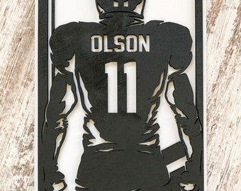 May include: Black silhouette of a football player with the name "OLSON" and the number "11" on the back of the jersey. The silhouette is cut out of wood and framed in a white frame.
