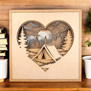 May include: Wooden wall art featuring a heart-shaped design with a camping scene. The artwork includes a tent, trees, and mountains, all intricately cut out. The frame is square and made of light-colored wood.
