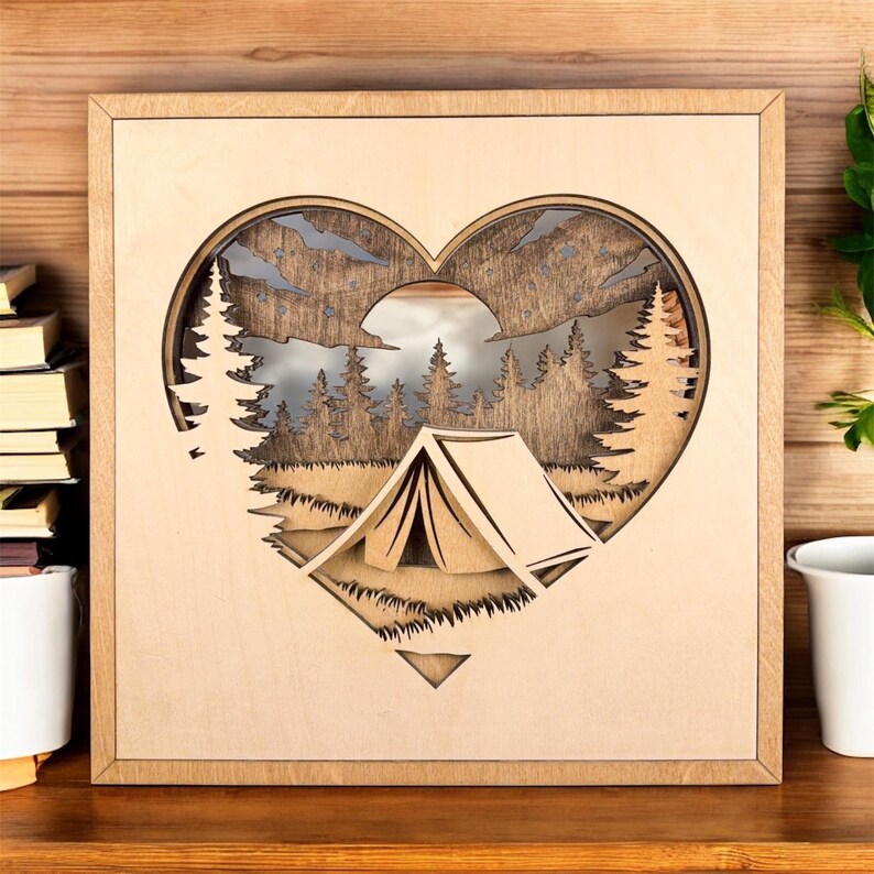 May include: Wooden wall art featuring a heart-shaped design with a camping scene. The artwork includes a tent, trees, and mountains, all intricately cut out. The frame is square and made of light-colored wood.
