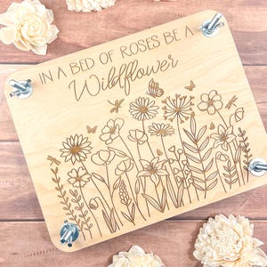 May include: A wooden craft board with the text "In a bed of roses be a wildflower" engraved on it. The board is decorated with a floral design and has four metal clamps on the sides.