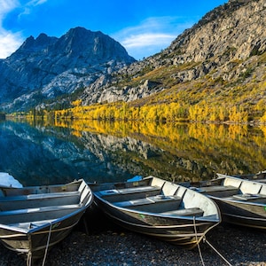 May include: A row of five aluminum boats are lined up on a rocky shore. The boats are all the same size and shape, and they are all facing the same direction. The boats are all empty, and they are all sitting on a bed of small rocks. The water is calm and still, and the sky is blue and clear. The mountains in the background are covered in trees, and the trees are all changing color. The water reflects the colors of the trees, creating a beautiful scene.