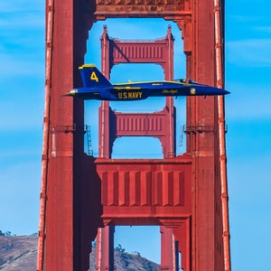 Może przedstawiać: Niebieski i żółty samolot myśliwski przelatuje przez most Golden Gate w San Francisco w Kalifornii. Samolot ma numer "4" i napis "U.S. NAVY" nadrukowany na boku. Samolot przelatuje przez czerwone wieże mostu.