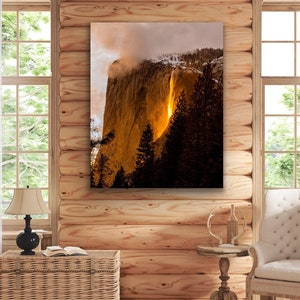 May include: A photograph of a waterfall cascading down a cliff face, with a golden glow emanating from the water. The cliff is a light brown color and the sky is a light blue with some clouds. The waterfall is surrounded by trees.