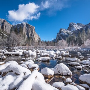 Könnte beinhalten: Eine schneebedeckte Landschaft mit einem Fluss, der durch sie fließt. Der Fluss ist von großen, schneebedeckten Felsen umgeben. Die Berge im Hintergrund sind ebenfalls schneebedeckt. Der Himmel ist blau und es gibt ein paar Wolken.