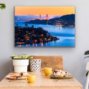 May include: A panoramic view of the Golden Gate Bridge at sunset, with the city lights twinkling in the distance. The bridge is a vibrant red against the orange and blue sky, with the water reflecting the colors.