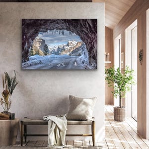 K&ouml;nnte beinhalten: Gerahmtes Landschaftsfoto, das eine schneebedeckte Stra&szlig;e durch einen Bergpass zeigt. Der Blick wird durch einen dunklen, felsigen Bogen mit Eiszapfen gerahmt. Der Himmel ist bedeckt, und die Berge sind in der Ferne zu sehen.