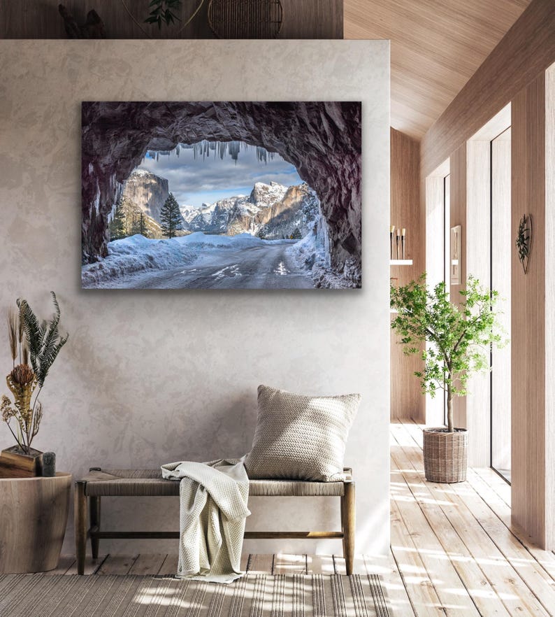 K&ouml;nnte beinhalten: Gerahmtes Landschaftsfoto, das eine schneebedeckte Stra&szlig;e durch einen Bergpass zeigt. Der Blick wird durch einen dunklen, felsigen Bogen mit Eiszapfen gerahmt. Der Himmel ist bedeckt, und die Berge sind in der Ferne zu sehen.