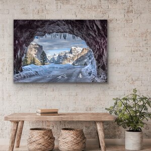 K&ouml;nnte beinhalten: Gerahmte Landschaftsfotografie einer schneebedeckten Berglandschaft, die durch eine H&ouml;hlen&ouml;ffnung betrachtet wird. Das Bild zeigt eine Stra&szlig;e, schneebedeckte Berge und Eiszapfen, die von der H&ouml;hlendecke h&auml;ngen. Das Foto wird &uuml;ber einer Holzbank mit zwei geflochtenen K&ouml;rben angezeigt.