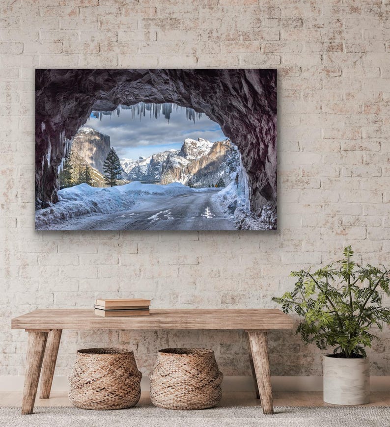 K&ouml;nnte beinhalten: Gerahmte Landschaftsfotografie einer schneebedeckten Berglandschaft, die durch eine H&ouml;hlen&ouml;ffnung betrachtet wird. Das Bild zeigt eine Stra&szlig;e, schneebedeckte Berge und Eiszapfen, die von der H&ouml;hlendecke h&auml;ngen. Das Foto wird &uuml;ber einer Holzbank mit zwei geflochtenen K&ouml;rben angezeigt.