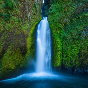 May include: A waterfall cascades down a moss-covered rock face, creating a pool of water at the bottom. The water is a vibrant blue, and the moss is a lush green.