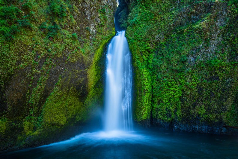 May include: A waterfall cascades down a moss-covered rock face, creating a pool of water at the bottom. The water is a vibrant blue, and the moss is a lush green.