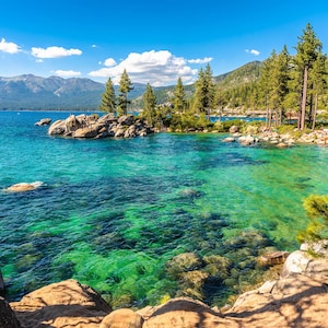 May include: A scenic view of a lake with clear, turquoise water and rocky shores. Lush green trees line the shoreline, creating a picturesque landscape.