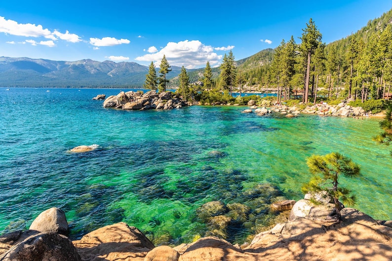 May include: A scenic view of a lake with clear, turquoise water and rocky shores. Lush green trees line the shoreline, creating a picturesque landscape.