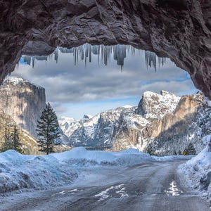 K&ouml;nnte beinhalten: Eine malerische Winterlandschaft, die durch einen Felsentunnel betrachtet wird. Schneebedeckte Berge und eine Stra&szlig;e sind zu sehen, mit Eiszapfen, die von der Tunneldecke h&auml;ngen. Der Himmel ist bew&ouml;lkt und erzeugt eine kalte, isolierte Atmosph&auml;re.