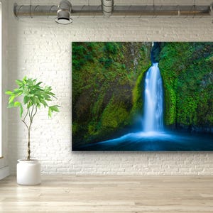 K&ouml;nnte beinhalten: Ein Foto eines Wasserfalls, der einen mit Moos bewachsenen Felsen hinunterst&uuml;rzt. Das Wasser ist leuchtend blau und die umliegende Vegetation ist ein sattes Gr&uuml;n.