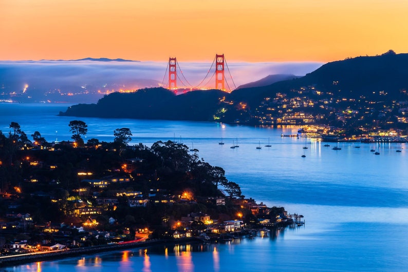 May include: A panoramic view of the Golden Gate Bridge at sunset, with the bridge illuminated in red and the surrounding water a deep blue. The city lights twinkle in the distance, and the fog rolls in over the hills.