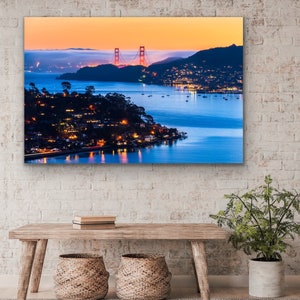 May include: A scenic photograph of the Golden Gate Bridge in San Francisco, California. The bridge is silhouetted against a vibrant orange and pink sunset sky. The water below the bridge is a deep blue, and the city lights twinkle in the distance.
