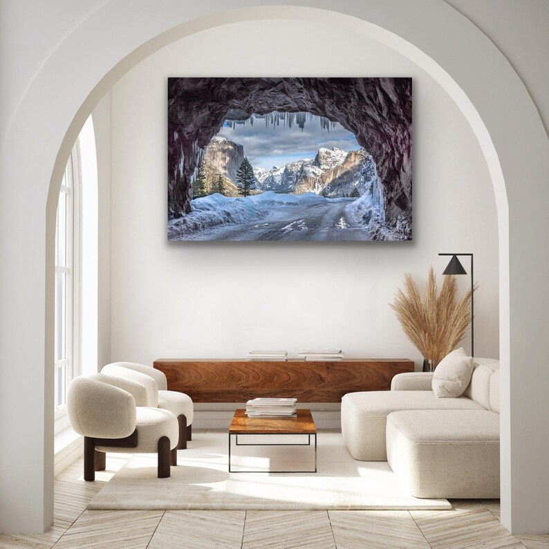 K&ouml;nnte beinhalten: Ein gerahmter Landschaftsdruck zeigt eine Winterlandschaft, die durch eine H&ouml;hlen&ouml;ffnung betrachtet wird. Das Bild zeigt schneebedeckte Berge, eine Stra&szlig;e und einen bew&ouml;lkten Himmel. Das Innere der H&ouml;hle ist dunkel, mit Eiszapfen, die von der Decke h&auml;ngen. Die Farbpalette ist k&uuml;hl, mit Wei&szlig;, Blau und Grau.