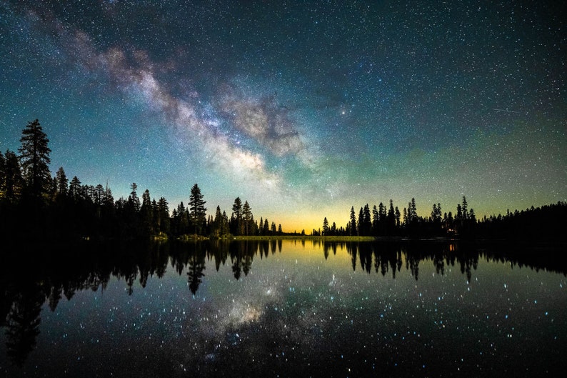 May include: A night sky filled with stars, including the Milky Way, reflected in a still lake. Silhouettes of trees line the shore.