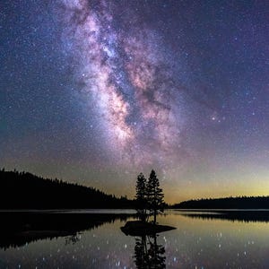 May include: A starry night sky with the Milky Way galaxy visible above a still lake. Two trees stand on a small island in the middle of the lake, their reflections mirrored in the water.