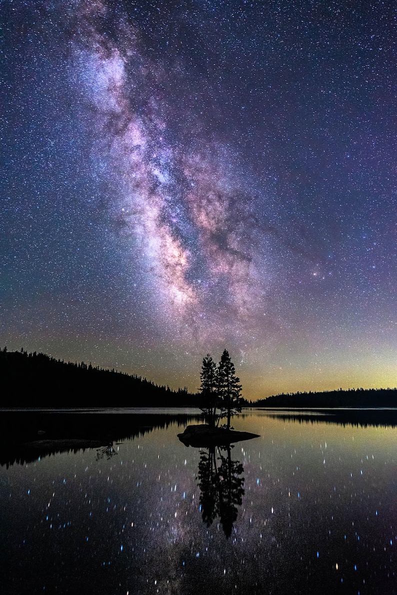 May include: A starry night sky with the Milky Way galaxy visible above a still lake. Two trees stand on a small island in the middle of the lake, their reflections mirrored in the water.
