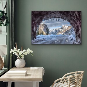 K&ouml;nnte beinhalten: Gerahmtes Landschaftsfoto einer verschneiten Berglandschaft. Das Bild wird durch eine dunkle H&ouml;hlen&ouml;ffnung mit Eiszapfen gerahmt. Eine Stra&szlig;e f&uuml;hrt durch das schneebedeckte Tal zu den Bergen.