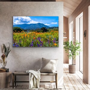 Puede incluir: Una fotograf&iacute;a de paisaje vibrante que presenta un campo de flores silvestres naranjas y moradas en primer plano, con una cordillera en el fondo. El cielo es azul con nubes blancas.