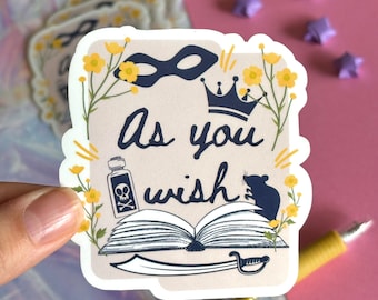 May include: A white sticker with a beige background featuring the text "As you wish" in black cursive script. The sticker also includes a black eye mask, a crown, a book, a sword, a bottle with a skull and crossbones, a rat, and yellow flowers.