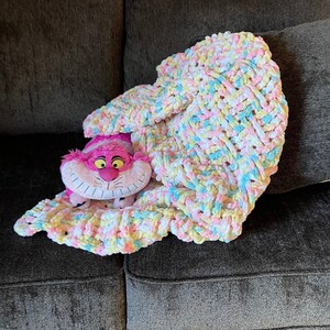 May include: A colorful crocheted blanket in shades of pink, yellow, and blue is draped over a couch. A plush pink Cheshire Cat toy is peeking out from under the blanket.