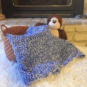 May include: A blue and white crocheted baby blanket is draped over a woven basket and a stuffed monkey toy. The blanket is soft and fluffy, perfect for a baby's nursery.