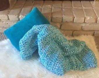 Könnte beinhalten: Eine tealfarbene und weiße Häkeldecke mit einem strukturierten Muster. Die Decke ist gefaltet und über einen weißen Kunstfellteppich gelegt.