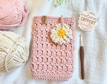 Pode incluir: Capa de telefone de crochê rosa com uma flor de crochê branca e uma etiqueta de madeira que diz "Bookish". Há um gancho de crochê e duas bolas de fio de fundo. A imagem também inclui um adesivo que diz "emotional support kindle".
