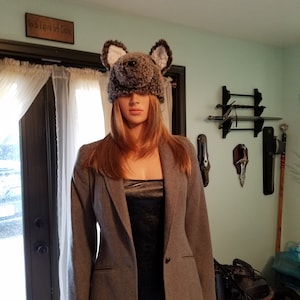 May include: A gray blazer with a single button closure. The blazer is worn over a black dress. The person is wearing a gray furry hat with bear ears.