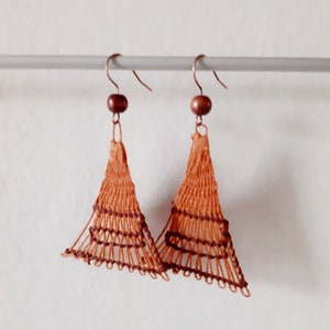 Puede incluir: Un par de pendientes triangulares tejidos de color marrón y naranja con cuentas de madera marrón en la parte superior.
