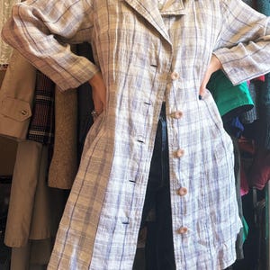 May include: A long-sleeved, button-down shirt featuring a light beige and blue plaid pattern. The shirt has a classic collar, a straight hemline, and five round, light-colored buttons running down the center front of the garment.