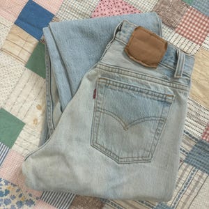 Puede incluir: Vaqueros de mezclilla azul claro con un parche de cuero marrón en la parte trasera. Los vaqueros están doblados y colocados sobre un edredón de retazos con varios colores y patrones. Los vaqueros tienen un diseño clásico de cinco bolsillos.