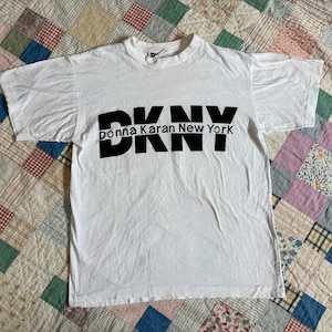 May include: White t-shirt with a black DKNY Donna Karan New York logo printed on the front.