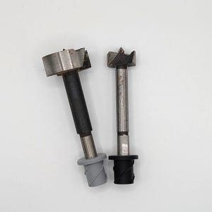May include: Two metal forstner drill bits. One has a black shaft and a grey base, the other a silver shaft and a black base. These drill bits are for woodworking, used to create flat-bottomed holes. The bits are made of metal.