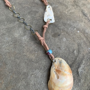 May include: A brown leather cord necklace with two shell pendants and a small blue and red bead. The larger shell pendant is a light brown color with a natural pattern.