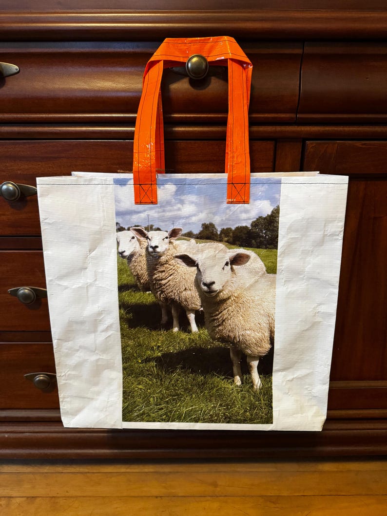 May include: White tote bag with a colorful image of three sheep in a green field. The bag has bright orange handles. The bag is made of a durable, possibly recycled material.