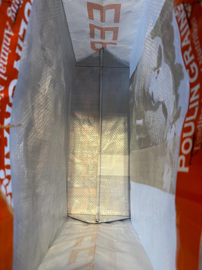May include: Inside view of a large, open, white and gray woven bag. The bag has a rectangular shape with a folded bottom. The word "FEED" is visible in orange letters on the top edge. The bag is likely for animal feed.