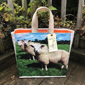 May include: A rectangular tote bag featuring a pastoral scene of three sheep in a green field under a blue sky. The bag has a cream-colored handle and a tag that reads "BagsForBasser". The interior trim is orange.