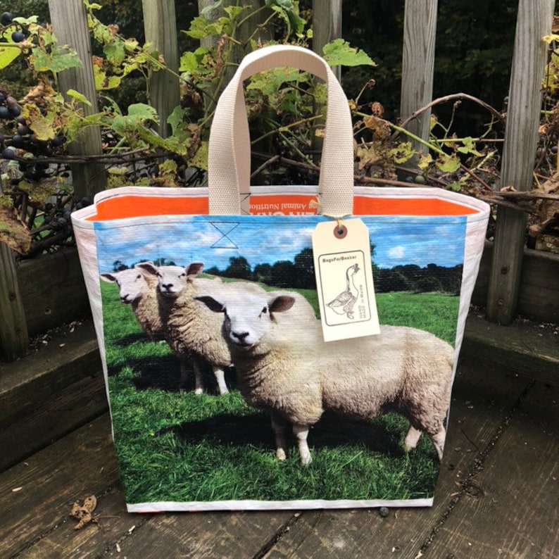 May include: A rectangular tote bag featuring a pastoral scene of three sheep in a green field under a blue sky. The bag has a cream-colored handle and a tag that reads "BagsForBasser". The interior trim is orange.