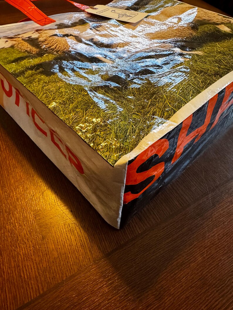 May include: A large, rectangular tote bag with a glossy, textured surface. The top of the bag features a pastoral scene of sheep grazing in a green field. The sides display the word "SALES" in red block letters against a black background.